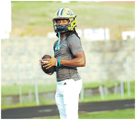 A person wearing a football helmet and uniform is holding a football on a field, appearing ready to throw. Description generated by AI