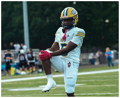 A person wearing a white football uniform with red gloves is standing on one leg on a football field with blurred players in the background. Description generated by AI
