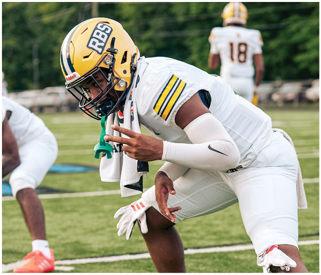 A person in white football gear and a yellow helmet is crouched on a grassy field, preparing for a play. Description generated by AI