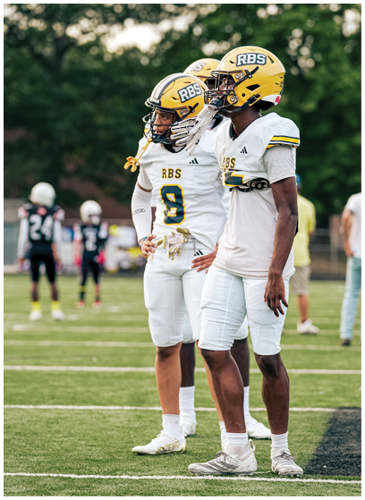 Two people wearing white football uniforms and yellow helmets stand on a grassy field during a game or practice. Description generated by AI
