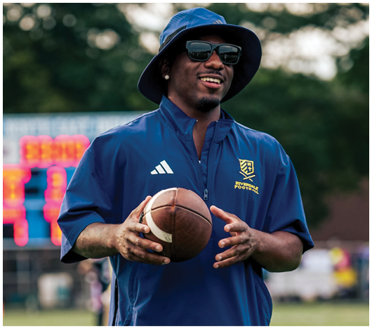 A person wearing sunglasses, a bucket hat, and a blue sports jacket is holding a football on a field with a blurred scoreboard in the background. Description generated by AI