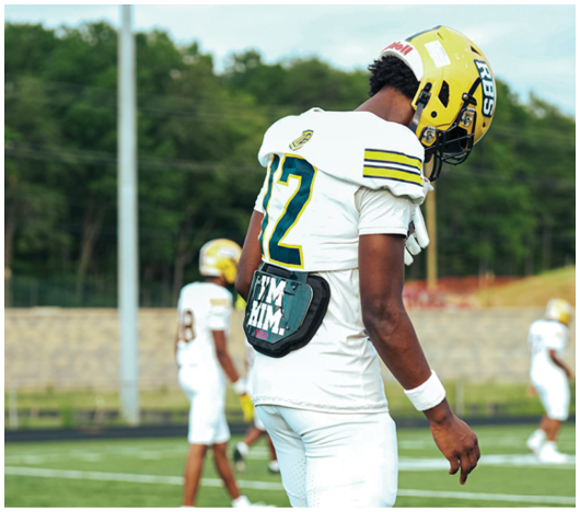 A person wearing a white football uniform with the number 12 and a yellow helmet stands on a football field with other players in the background. Description generated by AI