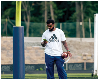 A person wearing a white Adidas shirt and sunglasses is holding a football and looking at a phone while standing near a yellow goalpost on a sports field. Description generated by AI