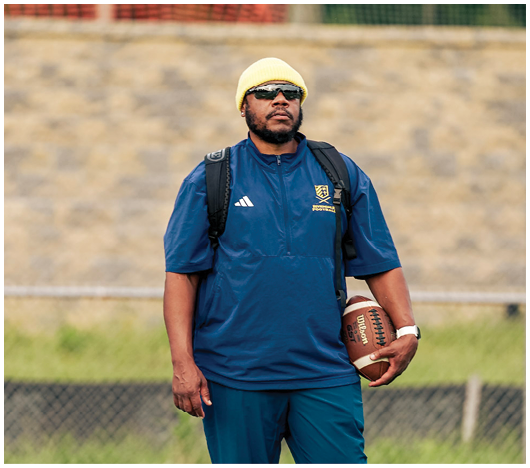 A person wearing a yellow beanie, dark sunglasses, a blue jacket, and a backpack stands outdoors with a blurred background of a stone wall and fencing. Description generated by AI