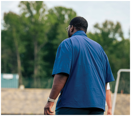An adult wearing a blue shirt and dark pants is standing on a sports field with a goalpost and net in the background. Description generated by AI