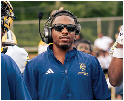 A person wearing sunglasses, a headset, and a blue sports jacket stands on a football field next to a person in a white football uniform holding a yellow helmet. Description generated by AI