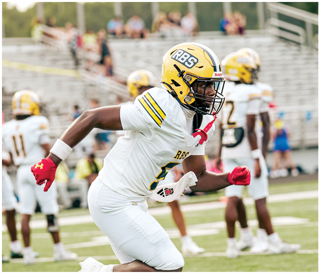 A person in a white football uniform with a yellow helmet and red gloves is running on a football field with other players and empty bleachers in the background. Description generated by AI