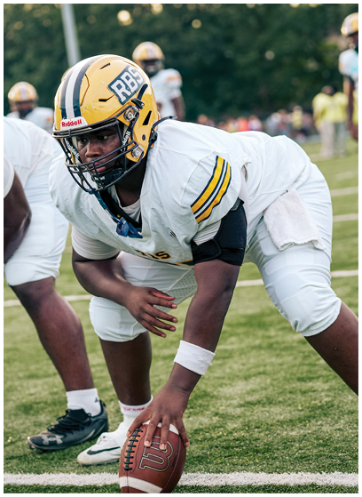 A person in a yellow helmet and white football uniform is crouched on a grassy field holding a football, preparing for a play. Description generated by AI