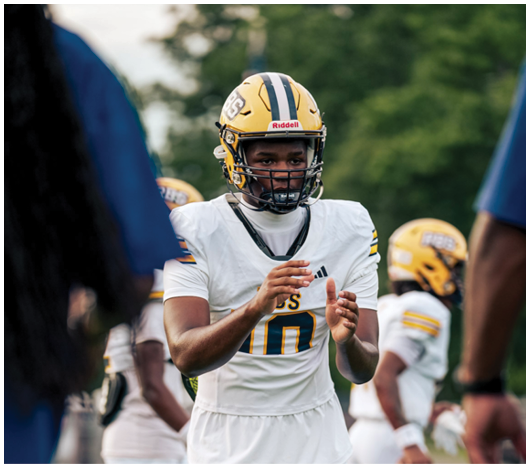 A person wearing a yellow football helmet and white jersey with the number 10 is clapping hands on a sports field with other players and a coach nearby. Description generated by AI