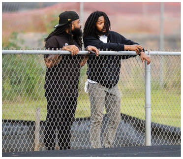 Two adults with long hair are leaning on a chain-link fence outdoors, engaged in conversation. Description generated by AI