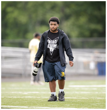 A person wearing a black jacket, graphic t-shirt, and blue shorts is walking on a sports field while holding a camera with a large lens. Description generated by AI