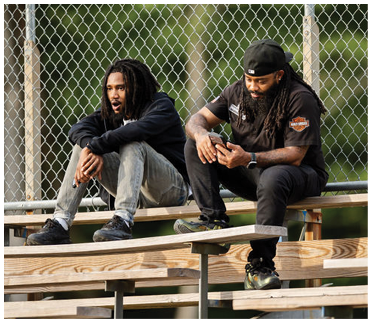 Two adults with long hair and casual clothing sit on wooden bleachers outdoors, one looking at a phone while the other looks ahead. Description generated by AI
