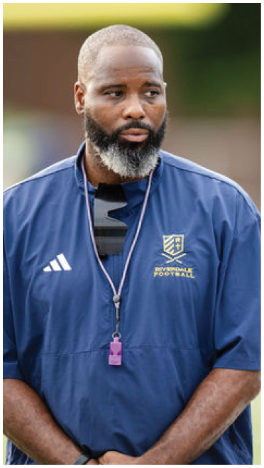 A person with a beard wearing a blue Riverdale Football jacket and a whistle around their neck stands outdoors with hands clasped. Description generated by AI