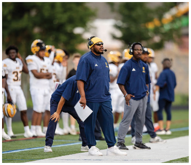 Adults wearing headsets and sportswear stand on the sidelines of a football field while players in white and yellow uniforms stand in the background. Description generated by AI