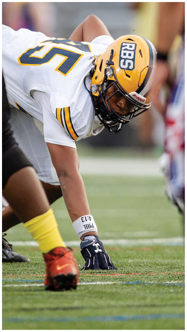 A person in a white football uniform and yellow helmet is crouched in a ready position on a football field facing another person wearing black pants and yellow socks. Description generated by AI