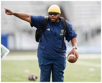 A person wearing a blue outfit is holding a football on a grassy field with another person partially visible in the background. Description generated by AI