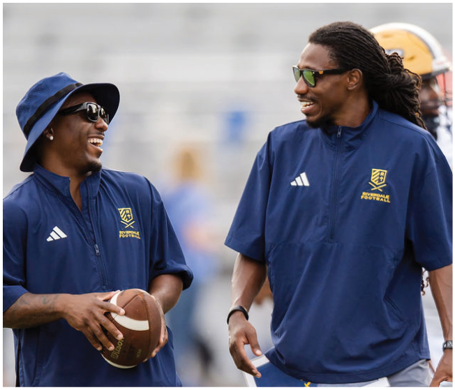 Two adults wearing matching sports jackets and sunglasses are smiling and holding a football while standing outdoors. Description generated by AI