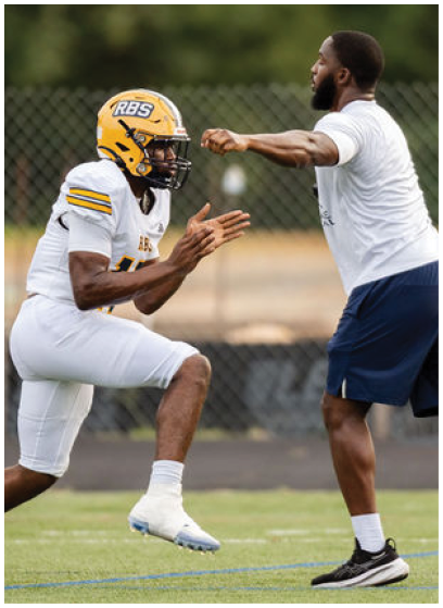 Two adults in yellow helmets and white football uniforms are on a field, with one standing and the other in a running stance holding a football. Description generated by AI