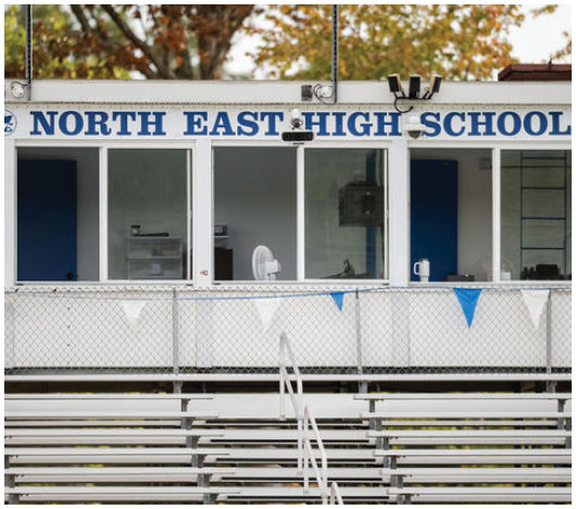 A person wearing a white shirt and dark pants is sitting alone on metal bleachers in front of a building labeled North East High School with blue doors and windows. Description generated by AI
