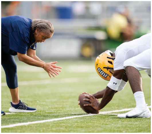A person in athletic clothing is bent forward with hands extended toward a football held by another person wearing a yellow helmet on a grassy sports field. Description generated by AI