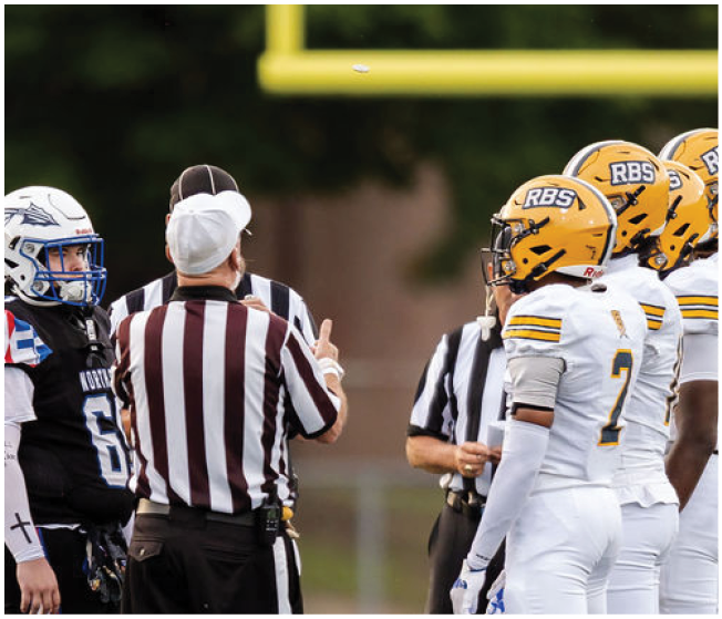 A group of people in football uniforms and referees are standing on a field under a goalpost, seemingly discussing or preparing for a play. Description generated by AI
