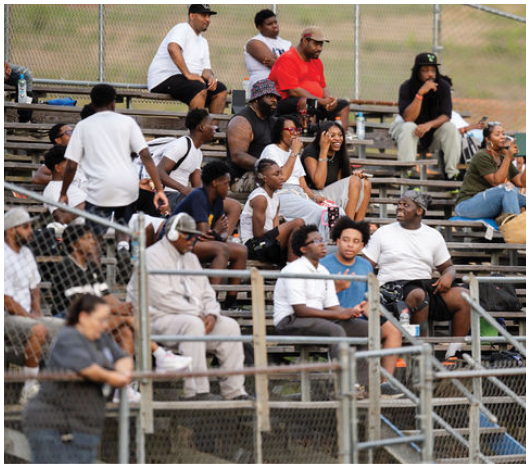 People sitting on bleachers outdoors, some talking, using phones, or watching an event. Description generated by AI