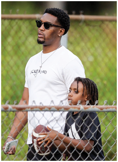 An adult wearing sunglasses and a white t-shirt stands behind a child with braided hair holding a football near a chain-link fence. Description generated by AI