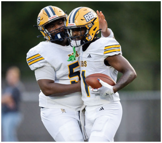 Two football players in white uniforms and yellow helmets are celebrating on the field, with one holding a football and the other raising a hand. Description generated by AI
