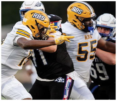 Two people in football uniforms are engaged in a play, with one player in a yellow helmet and white uniform reaching out toward another player in a black uniform. Description generated by AI
