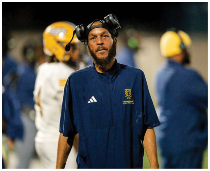 A person wearing a headset and a navy blue sports shirt with a logo stands on a football field with blurred players in helmets in the background. Description generated by AI