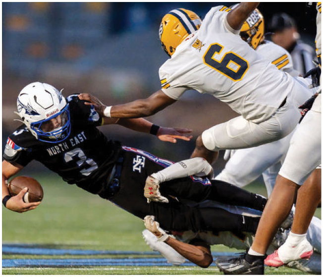 A person in a black football uniform is diving forward while holding a football as a person in a white and yellow uniform reaches out to tackle them on a grassy field. Description generated by AI