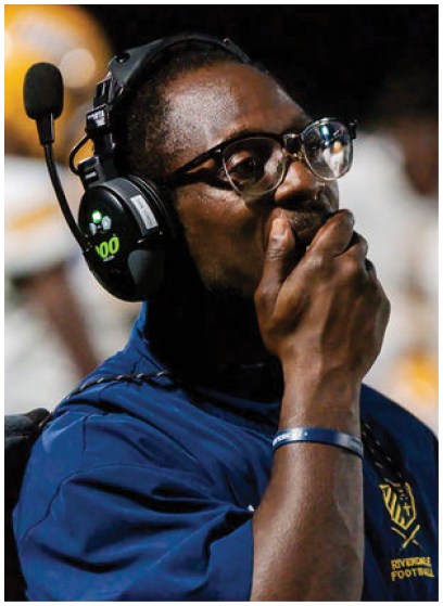 A person wearing glasses and a headset is speaking into a microphone while standing on a sports field with blurred players in the background. Description generated by AI
