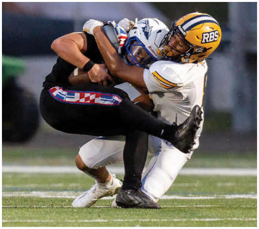 Two people in football uniforms are engaged in a tackle on a grassy field during a game. Description generated by AI