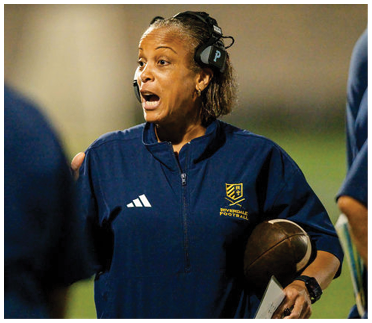 A person wearing a headset and a Riverside Football jacket is holding a football and appears to be speaking or giving instructions during a game. Description generated by AI