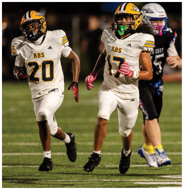 Two people in white football uniforms with yellow helmets and pink gloves are running on a field while a person in a black uniform with a white helmet follows behind them. Description generated by AI