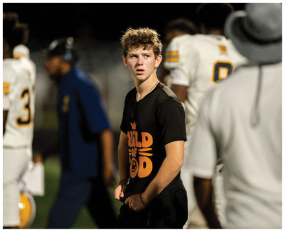 A person with curly hair wearing a black t-shirt with orange text stands on a sports field while several people in white sports uniforms and one in a blue outfit move in the background. Description generated by AI