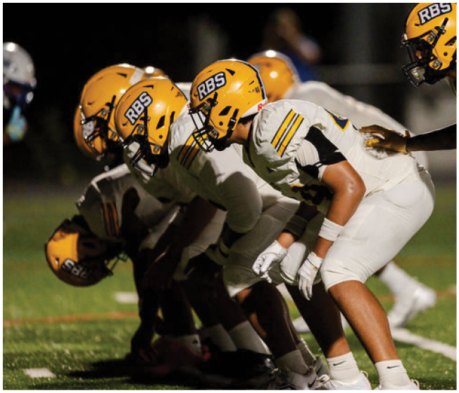 A group of football players in yellow helmets and white uniforms are lined up in a crouched position on a grassy field, preparing for a play. Description generated by AI