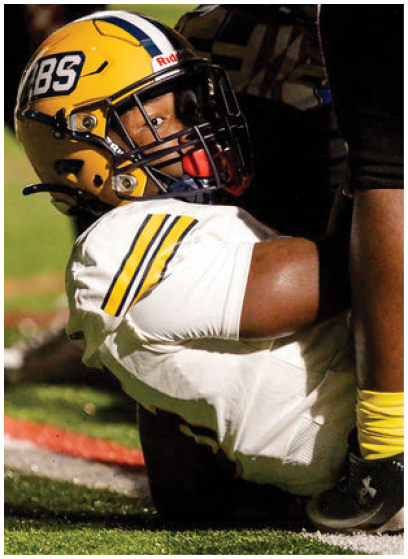 A person wearing a yellow football helmet and white jersey with yellow and black stripes is lying on the ground on a football field. Description generated by AI