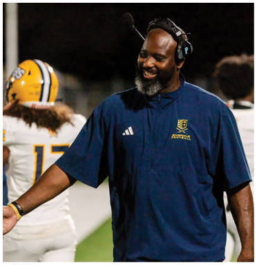 A person wearing a headset and a blue sports shirt is smiling and reaching out to shake hands with another person on a football field while a player in a yellow helmet and white uniform walks away in the background. Description generated by AI