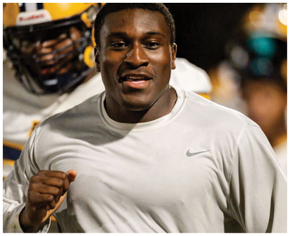 A person wearing a light gray Nike shirt is running with a determined expression while another person in football gear is visible in the background. Description generated by AI