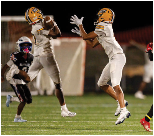 Two people in white football uniforms with yellow helmets are catching a football on a field while other players in dark uniforms are nearby. Description generated by AI