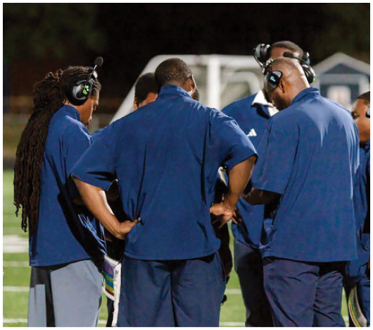 A group of people wearing blue shirts and headsets are standing together on a sports field, seemingly engaged in a discussion. Description generated by AI
