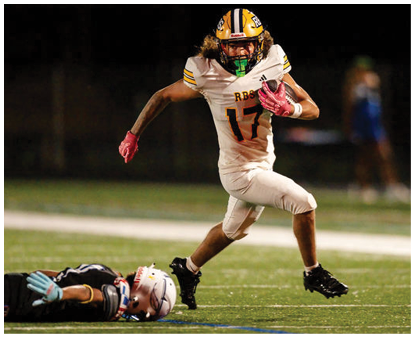A person wearing a yellow helmet and white football uniform with the number 17 is running on a football field while another person in a black uniform lies on the ground. Description generated by AI