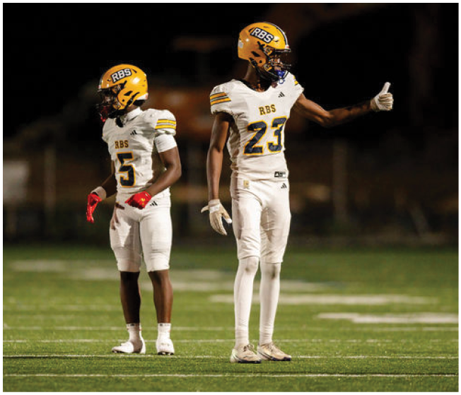 Two people in white football uniforms and yellow helmets stand on a field, with one giving a thumbs-up gesture. Description generated by AI