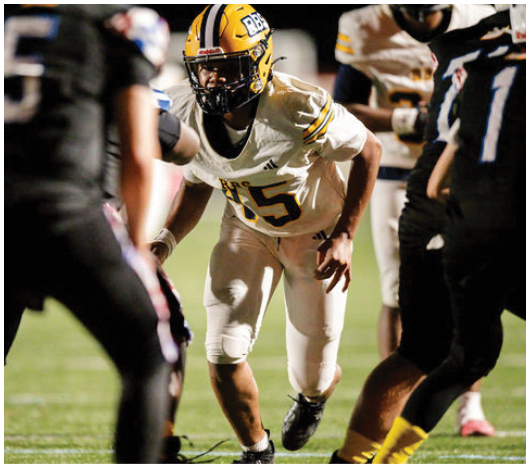 A person in a yellow and white football uniform is crouched and ready to move on a football field while two opposing players in black uniforms stand nearby. Description generated by AI