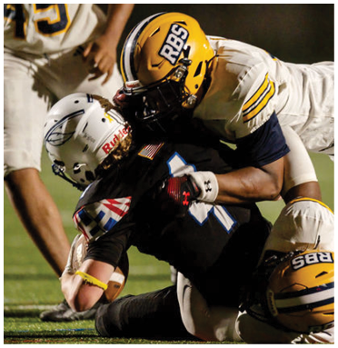 Two football players in white uniforms and yellow helmets tackle a player in a black uniform during a game on a grassy field. Description generated by AI