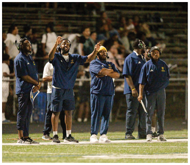 A group of adults wearing matching navy blue athletic outfits stand on a sports field with one person gesturing animatedly while others watch attentively. Description generated by AI
