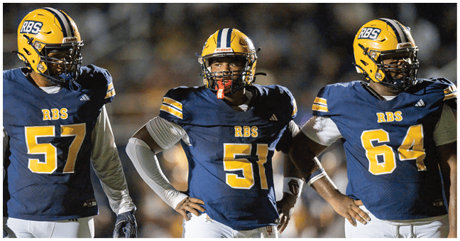 A group of football players in navy and white uniforms huddle together on a field near a scoreboard showing the first period and a score of zero. Description generated by AI