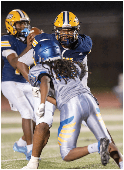 Two people wearing blue and yellow football uniforms and helmets stand on a field with one person holding a hand near their face and the other looking forward. Description generated by AI
