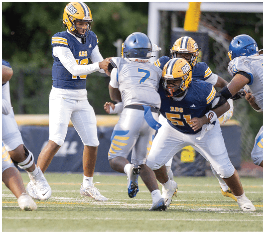 A person wearing a blue and yellow football uniform with the number 57 is running on a field with other players blurred in the background. Description generated by AI